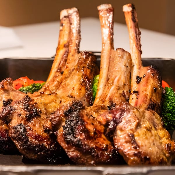 Four grilled lamb chops with visible bones, served with broccoli and red peppers.