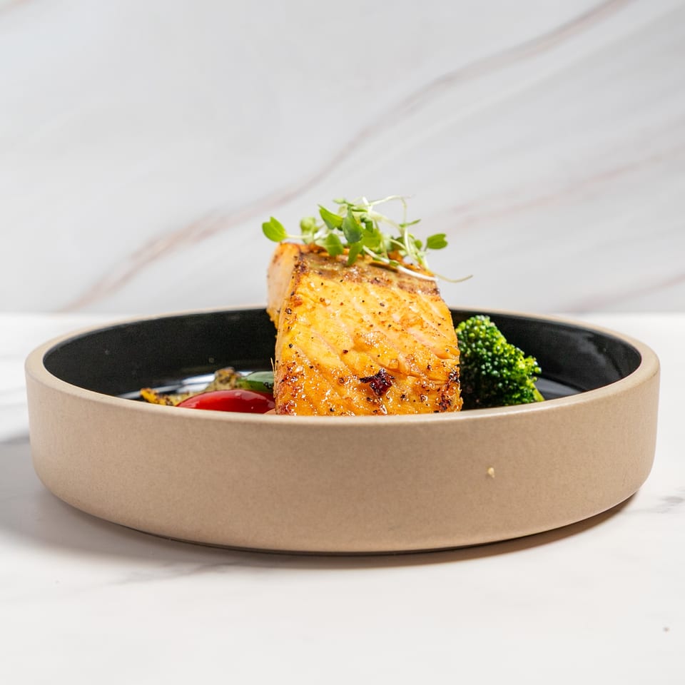 Golden-brown salmon fillet topped with microgreens, served with broccoli and red pepper in a bowl.