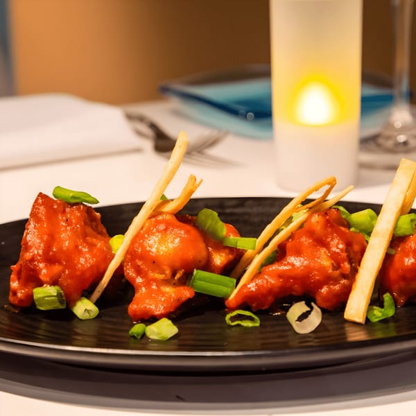 Indian chicken appetizer with red sauce, green onions, and crispy garnish on a black plate.